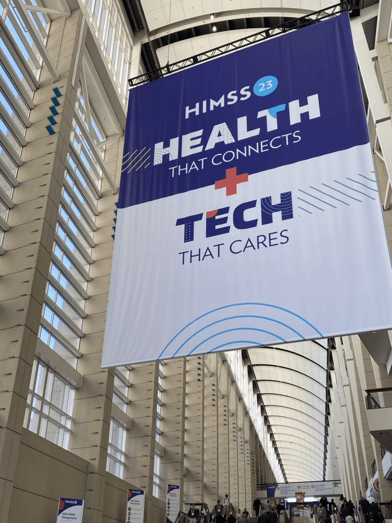 Photo of a large banner hanging in a long bright hallway which states HIMSS 23, Health that connects & Tech that cares