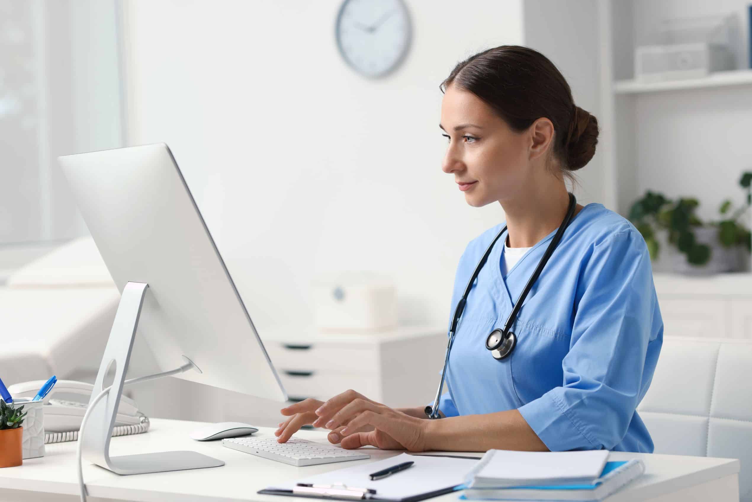 Female medical professional in light blue scrubs inputs data into a computer.