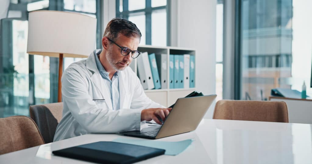Healthcare, laptop and report with doctor man at desk in office of hospital for diagnosis, planning or research. Computer, medical and mature medicine professional with pointing or reading in clinic.