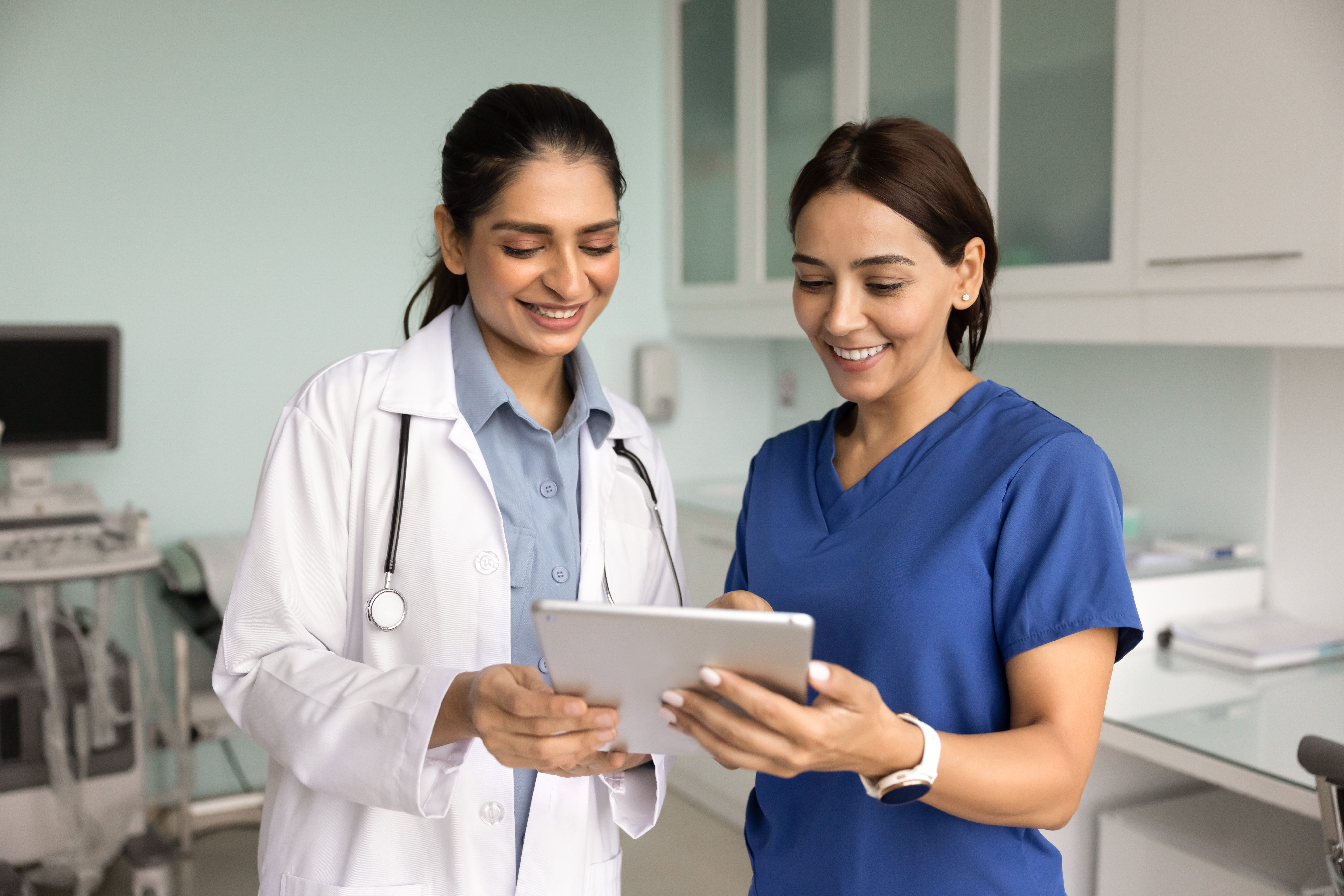 Positive diverse young medical colleagues discussing patient electronic records, exam results on tablet, looking and pointing at gadget, talking, smiling, laughing in clinic examination room
