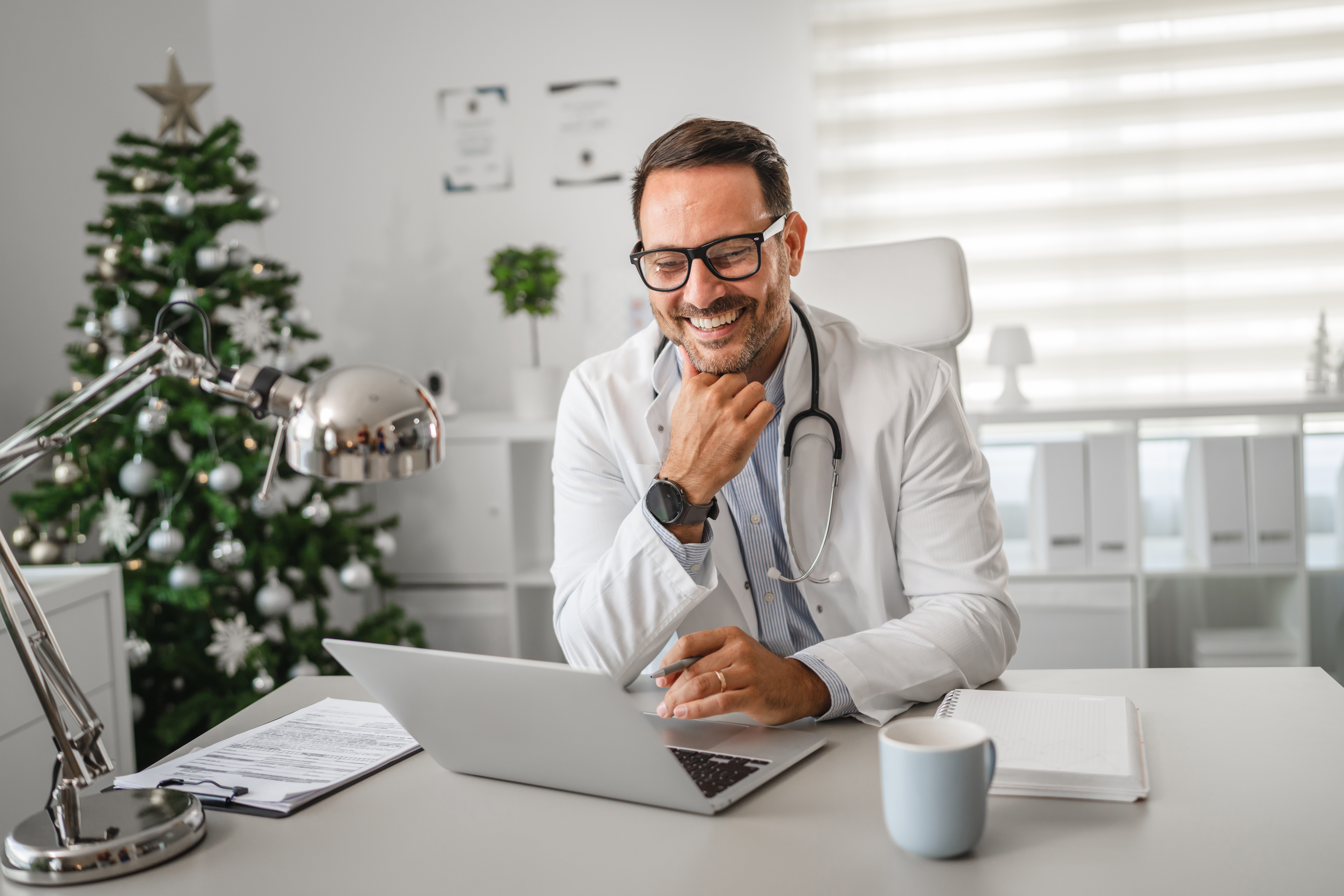 Smiling male doctor wearing lab coat and stethoscope using laptop at desk in medical office with decorated christmas tree, enjoying festive season while working
