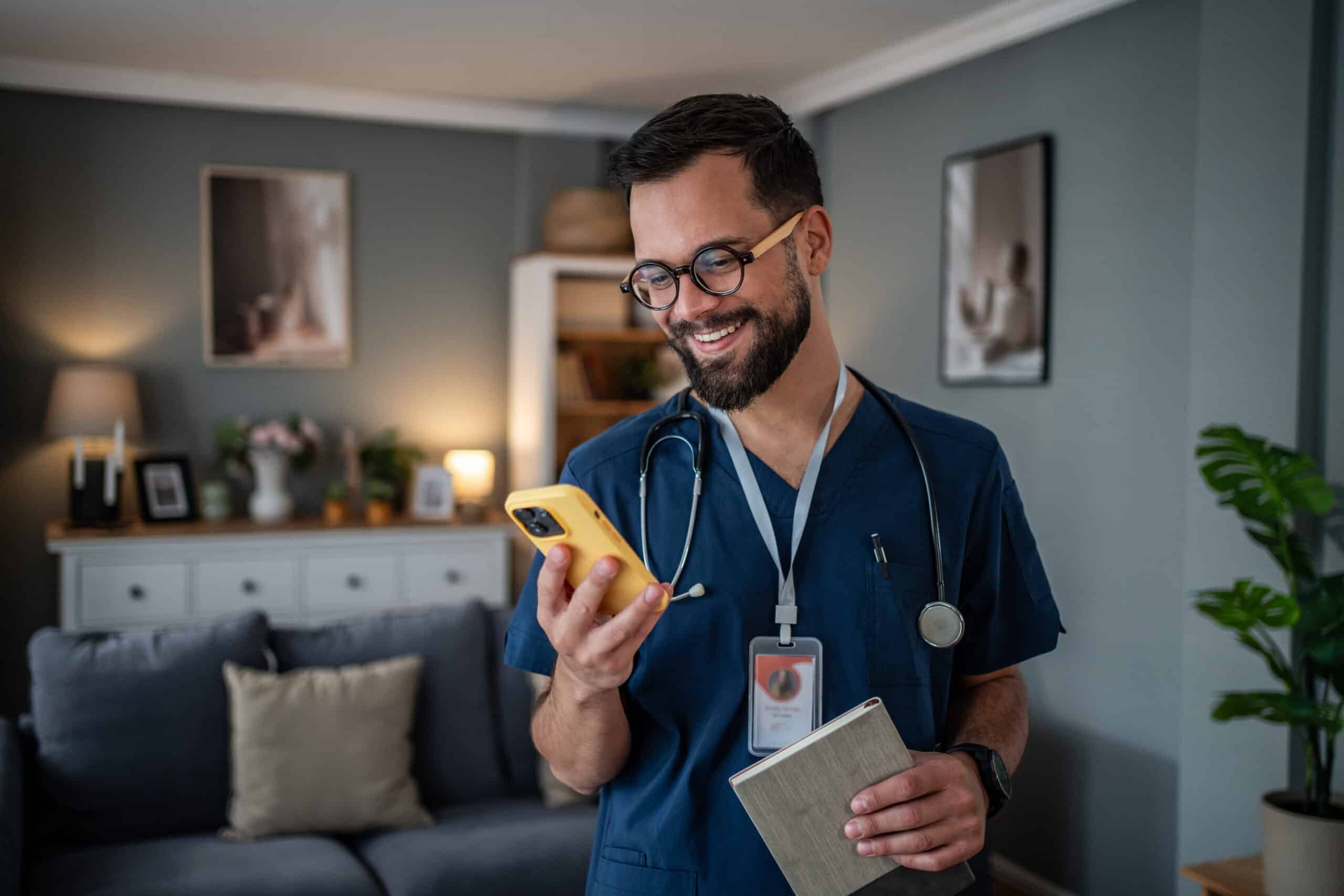 Young male doctor or nurse wearing medical scrubs and a stethoscope, smiling while holding a smartphone and a notebook, offering virtual consultation or home healthcare service