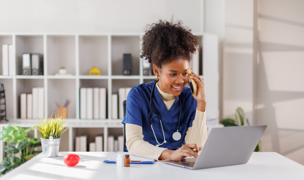 Happy friendly young woman doctor, nurse, medical worker, reception specialist in blue uniform with stethoscope sitting at desk, talking to patient on phone and checking schedule information on laptop