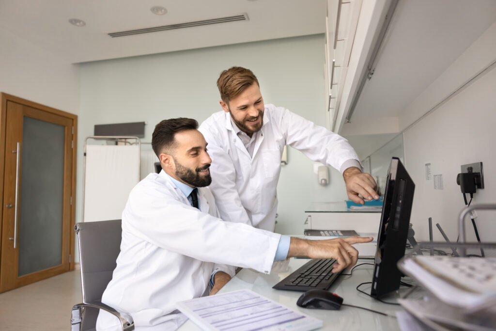 How AI Document Analysis Supports Smarter Clinical Workflows Digital healthcare achievement. Two young male doctors mentor and intern point at desktop monitor with smiles make diagnosis together review correct treatment result confirm positive patient progress
