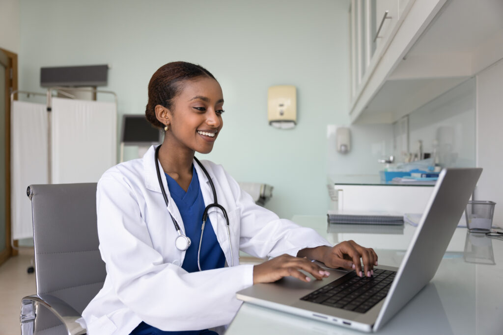 How AI Document Analysis Supports Smarter Clinical Workflows Happy young African general practitioner woman working at computer in medical office, typing report, records on laptop, smiling, chatting with patients, providing telehealth service