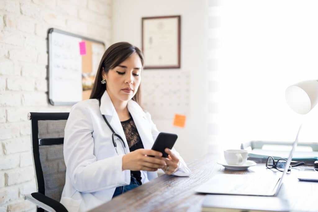 How Secure Patient Messaging Improves Care Coordination Portrait of young hispanic woman doctor sitting at her office desk and texting on mobile phone