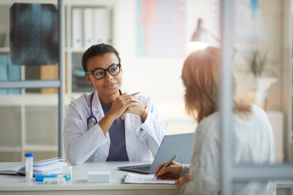 Young female doctor in white coat sitting at the table and listening to patient sitting in front of her at office