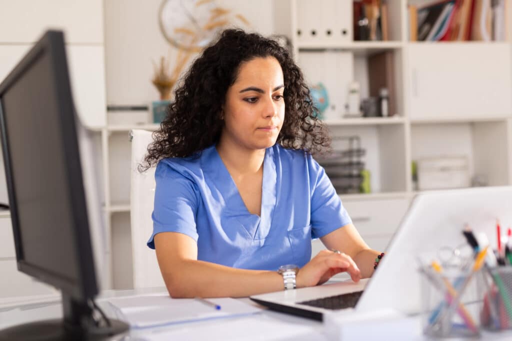 Young woman doctor assistant working in medical office using laptop computer and writing prescription