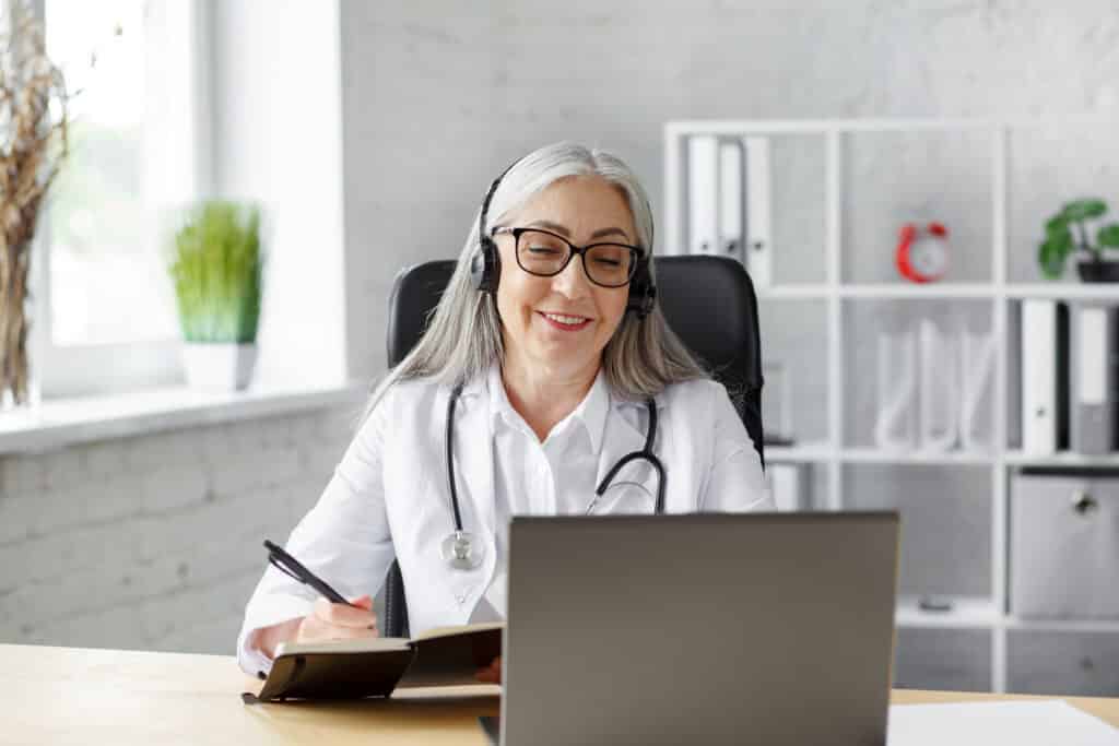 Why HIPAA-Compliant Telehealth Is Vital for Your Practice Portrait of senior grey-haired female doctor in her office using laptop for video chat with a patient. Online consultation with doctor for diagnoses and treatment recommendation. Telehealth concept.