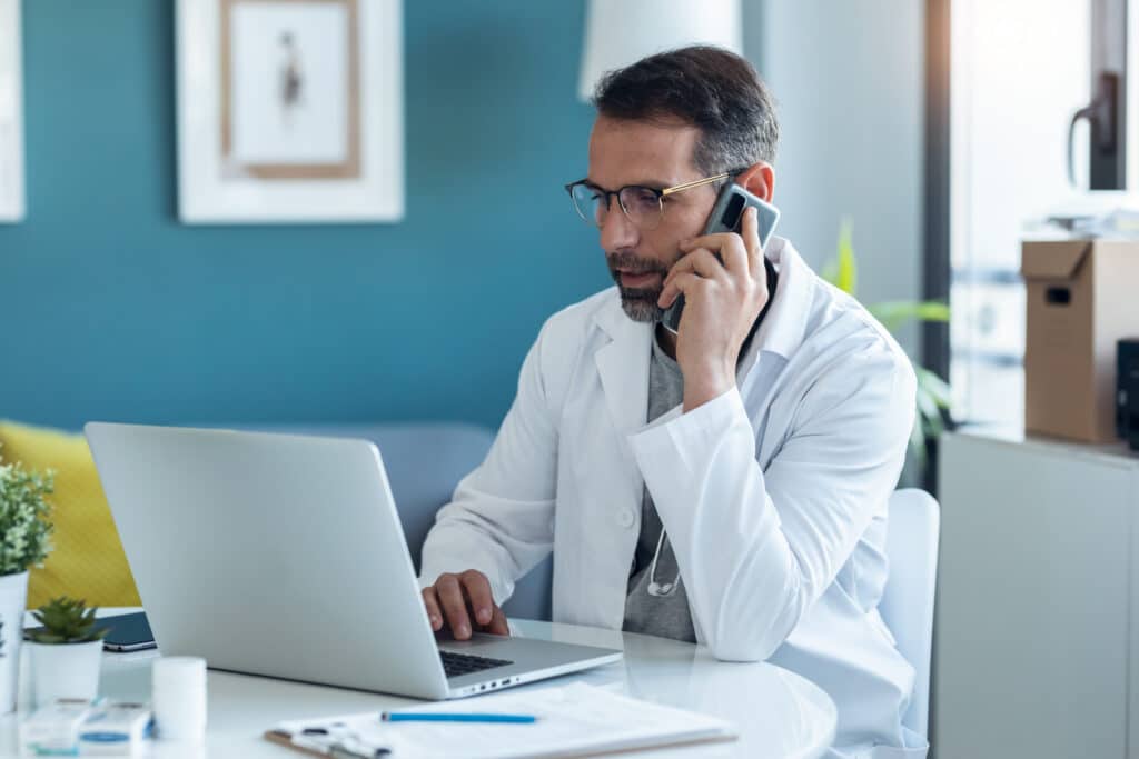 Why HIPAA-Compliant Telehealth Is Vital for Your Practice Shot of mature concentrated attractive male doctor working with her computer while talking with smartphone in medical consultation.