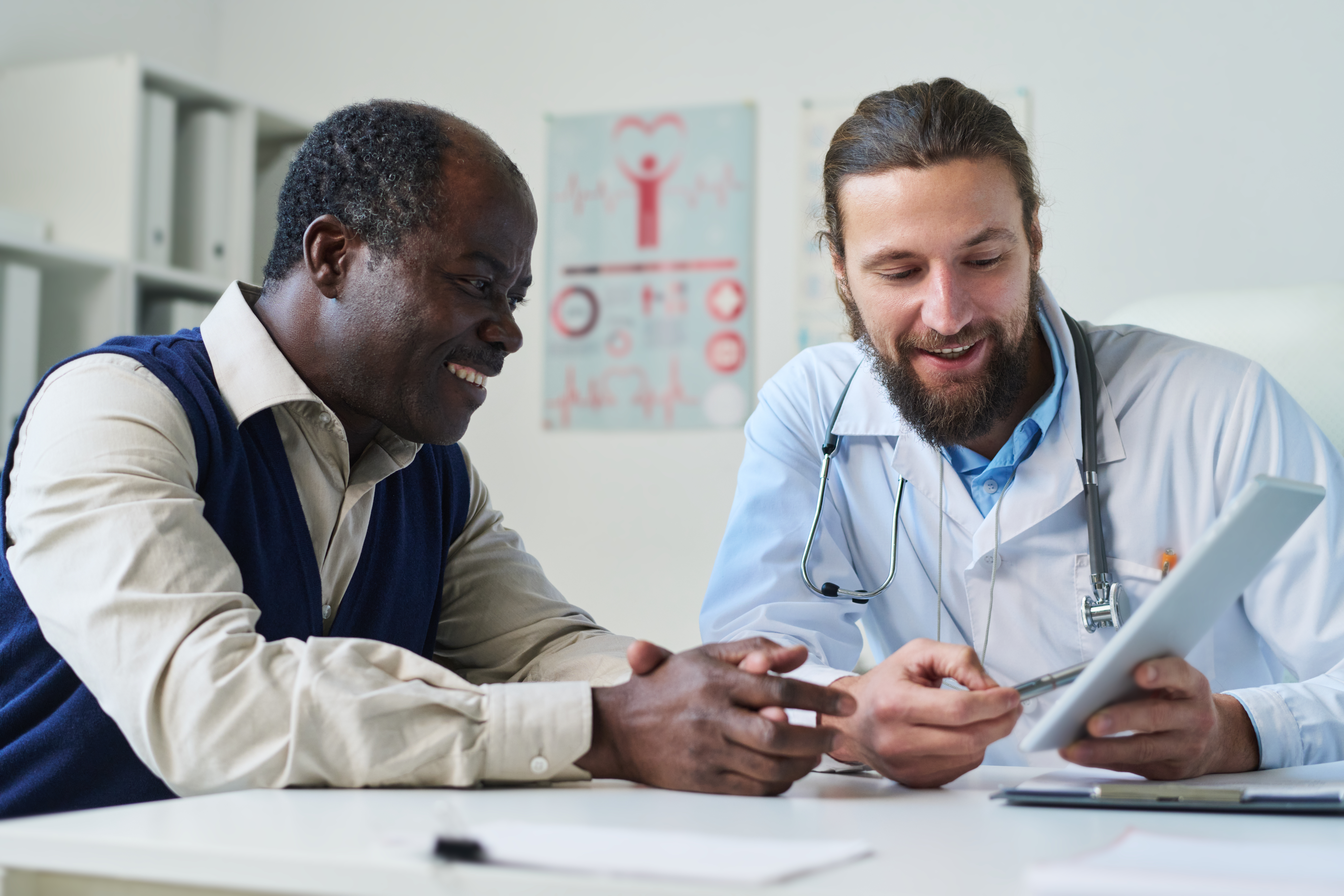 Young smiling doctor pointing at tablet screen with results of medical test and explaining diagnosis to senior African American male patient