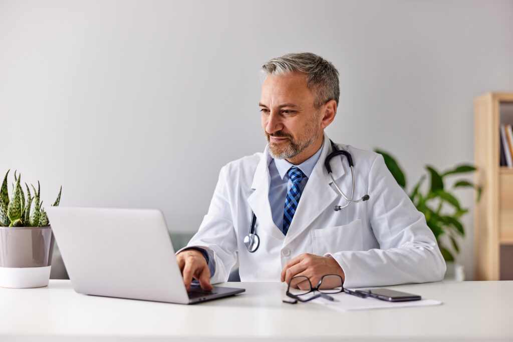 HIPAA-Compliant Fax vs. Generic Online eFax Platforms: What's the Difference? A senior male doctor typing on a laptop, sitting at the clinic, wearing a white coat.