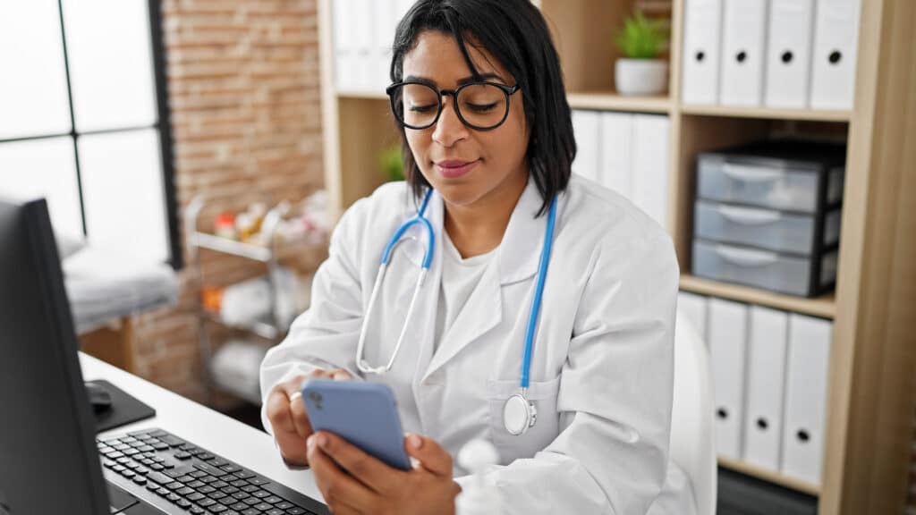 How AI Document Analysis Supports Smarter Clinical Workflows A hispanic woman doctor checks her smartphone in a well-lit hospital office, evoking a busy healthcare setting.