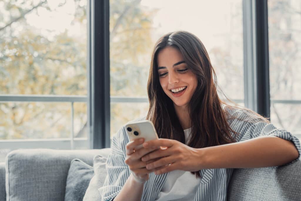 How Automated Appointment Reminder Software Reduces No-Shows Happy pleasant millennial woman relaxing on comfortable couch, holding smartphone in hands. Smiling young lady chatting in social networks, watching funny videos, using mobile applications at home.